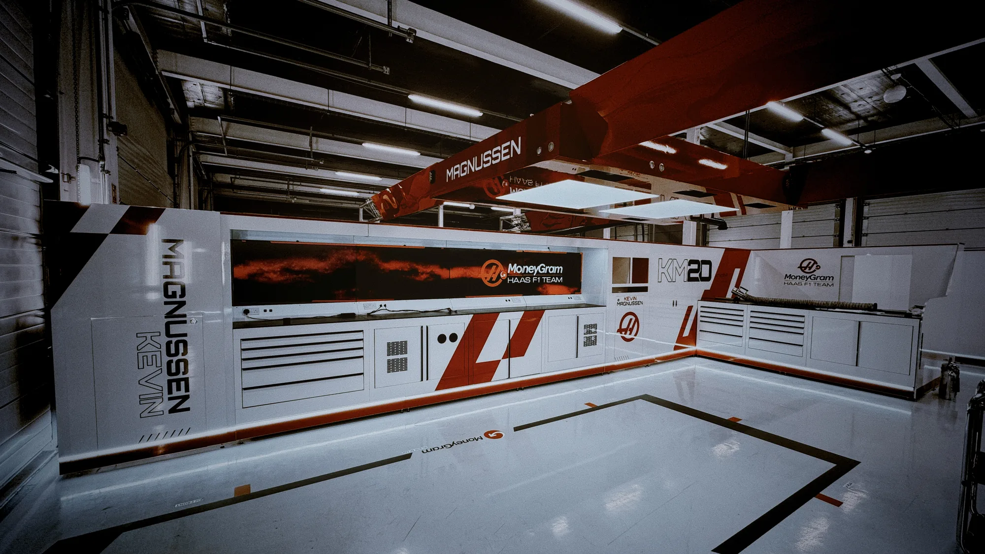 A Formula One pit garage fitted out with modular walling panels in white and red, with branded cabinetry and an overhead gantry.